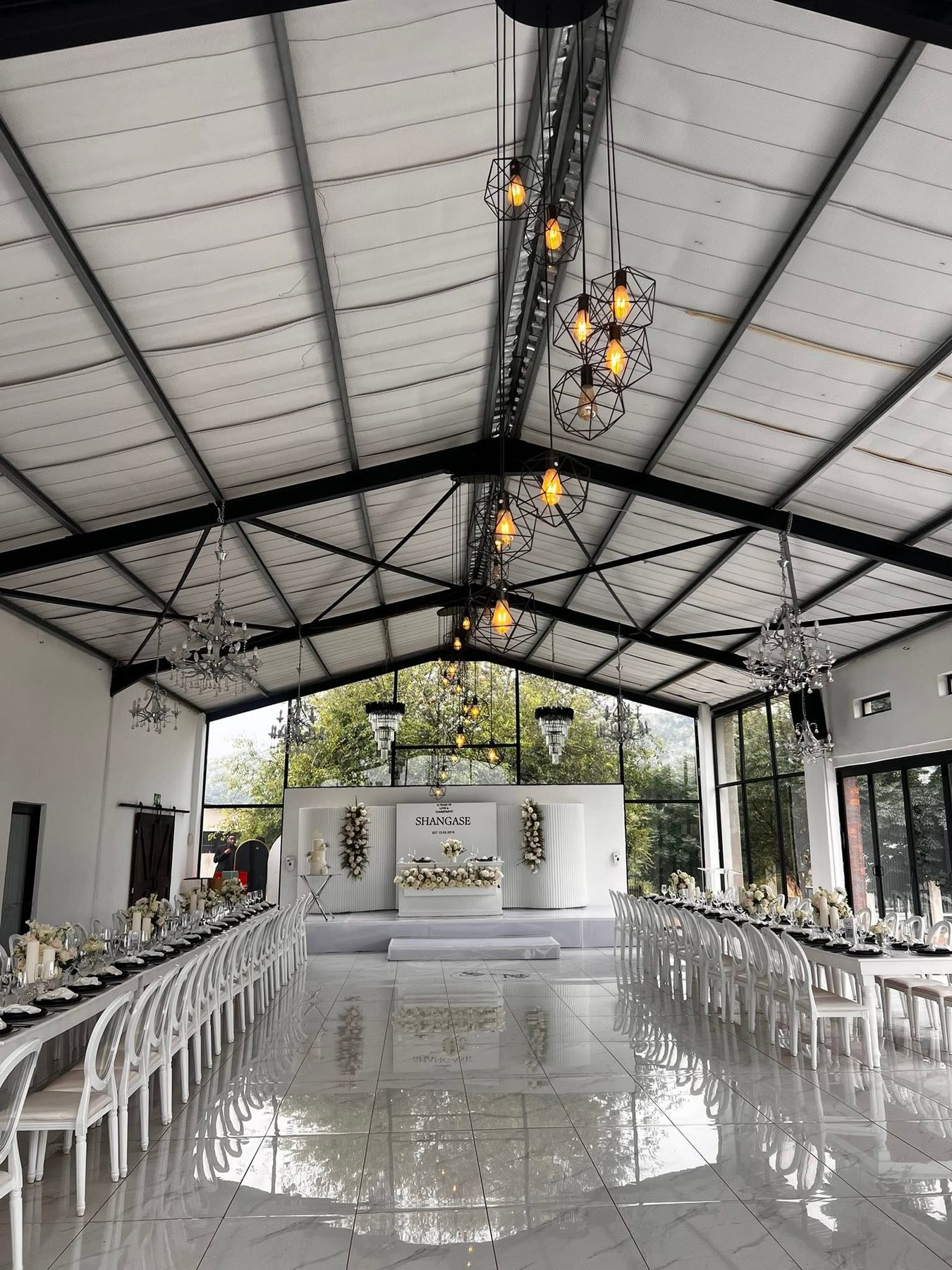 Glass pavilion set for a black-and-white reception, chandeliers overhead and long banquet tables either side